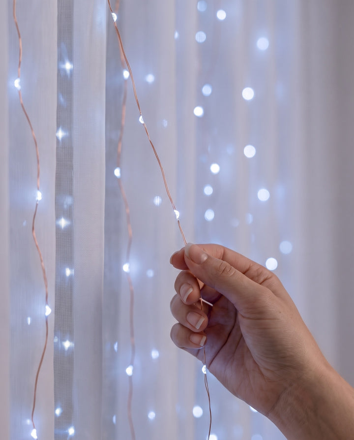 Close-up of a hand holding a strand of cool white LED fairy curtain lights on copper wire in front of a sheer curtain