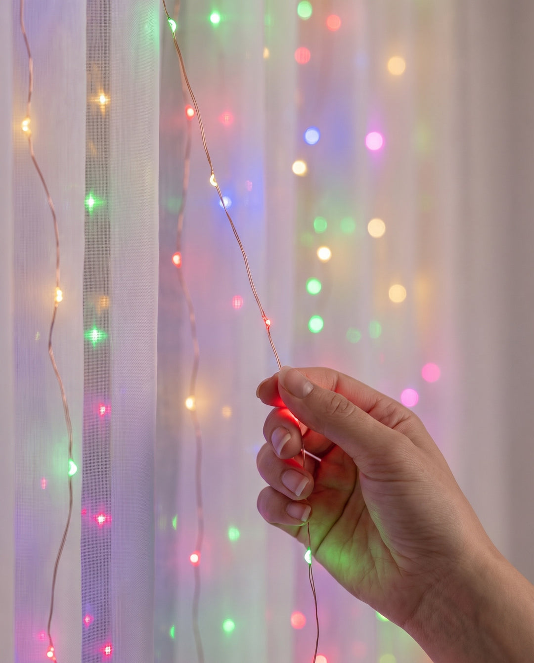 Close-up of a hand holding a strand of multicolor LED fairy curtain lights on copper wire in front of a sheer curtain