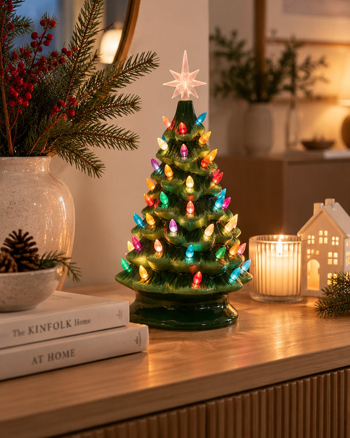 Ceramic Christmas tree glowing on a wooden console beside a candle and a small ceramic house