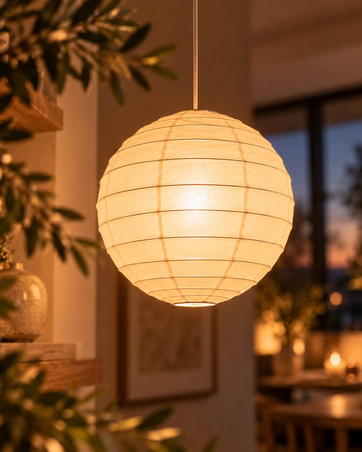 Close-up of a round paper lantern pendant lit among olive branches at dusk