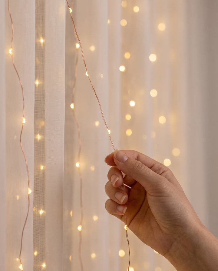 Close-up of a hand holding a strand of tiny warm white LEDs on fine copper wire in front of a sheer curtain