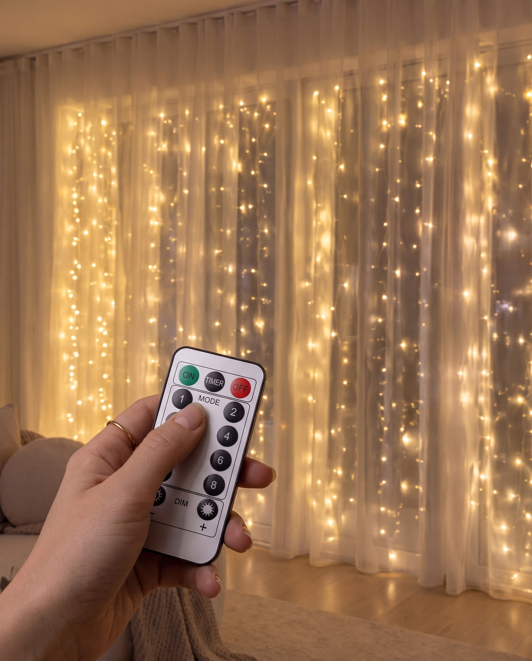 Hand holding the remote control in front of warm white LED fairy curtain lights behind sheer curtains