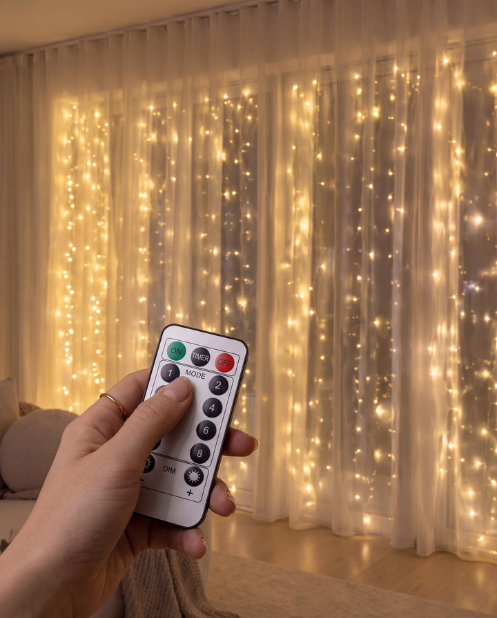 Hand holding the remote control in front of warm white LED fairy curtain lights behind sheer curtains