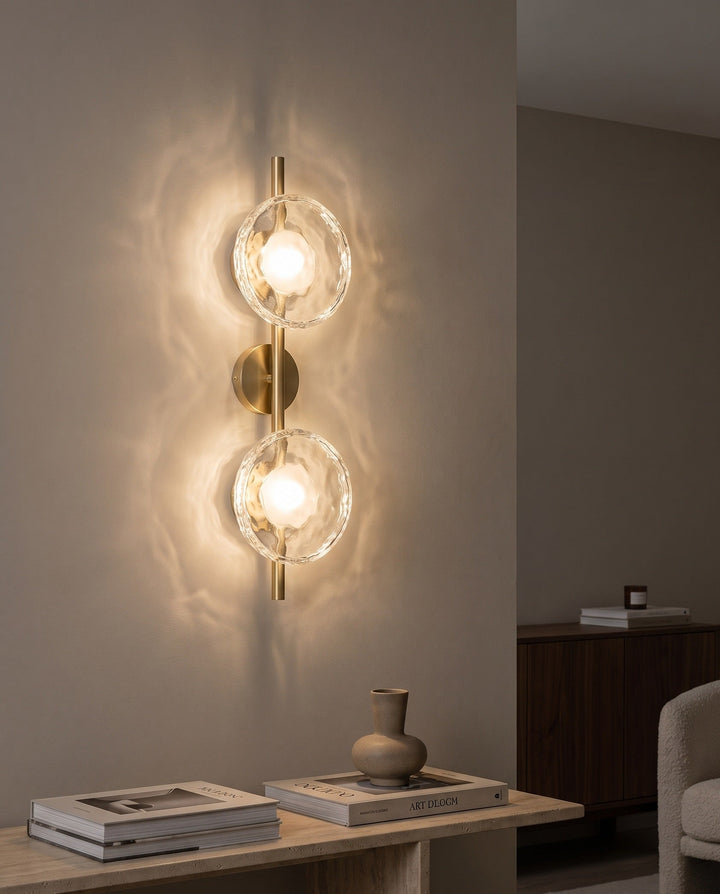 Orbe double head wall sconce in gold with clear glass glowing above a travertine console