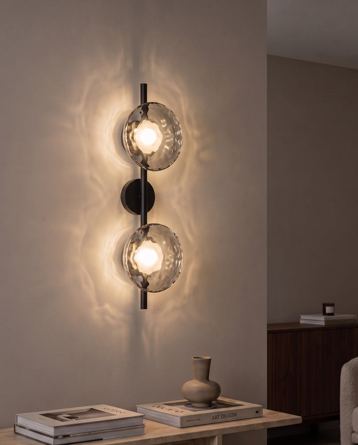 Orbe double head wall sconce in black with smoked glass glowing above a travertine console