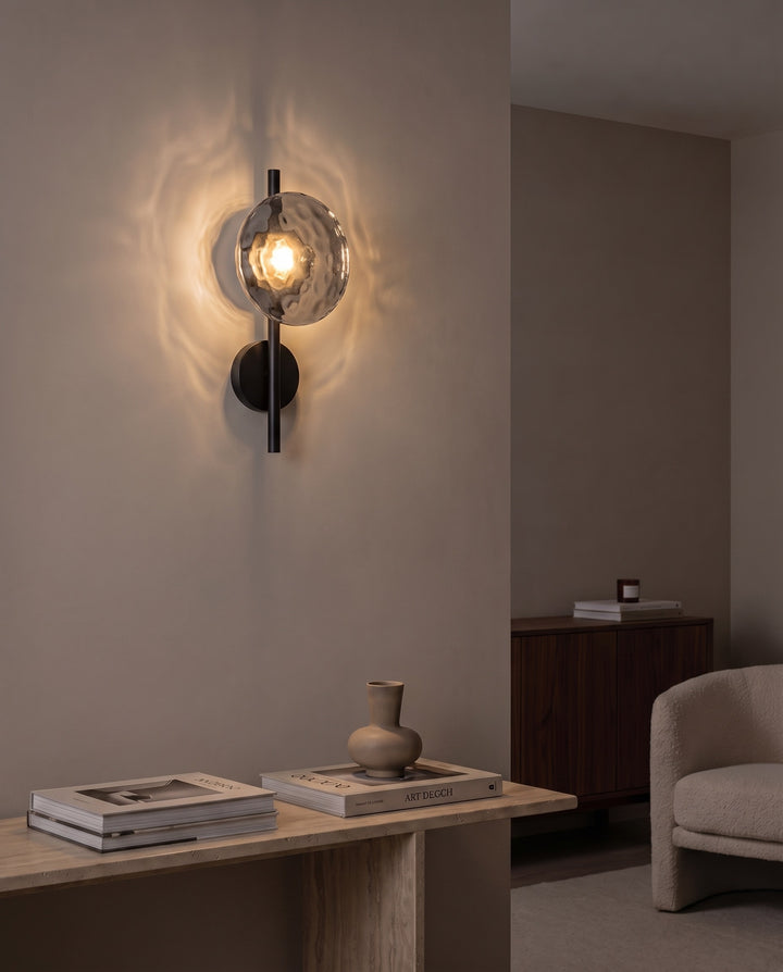 Orbe single head wall sconce in black with smoked glass glowing above a travertine console