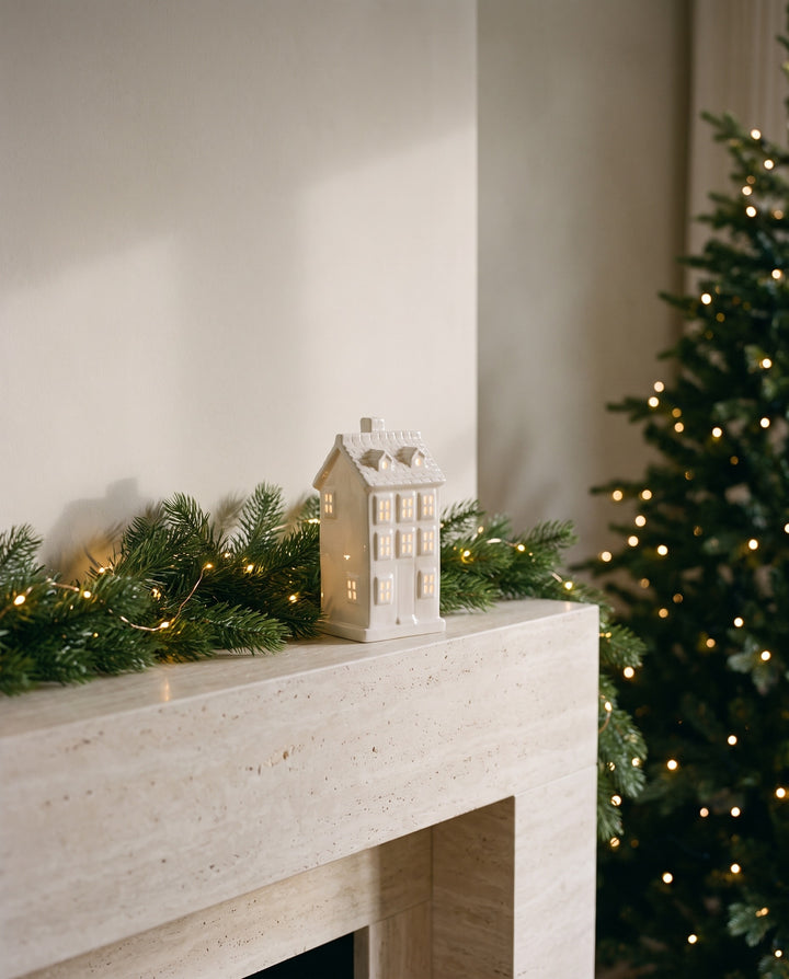 White ceramic Villa house with glowing windows on a travertine mantelpiece with a fir garland