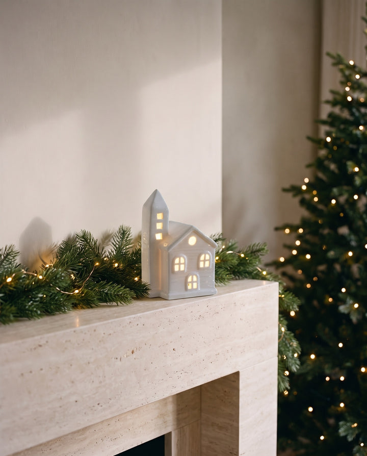 White ceramic house with a bell tower and glowing windows on a mantelpiece at Christmas