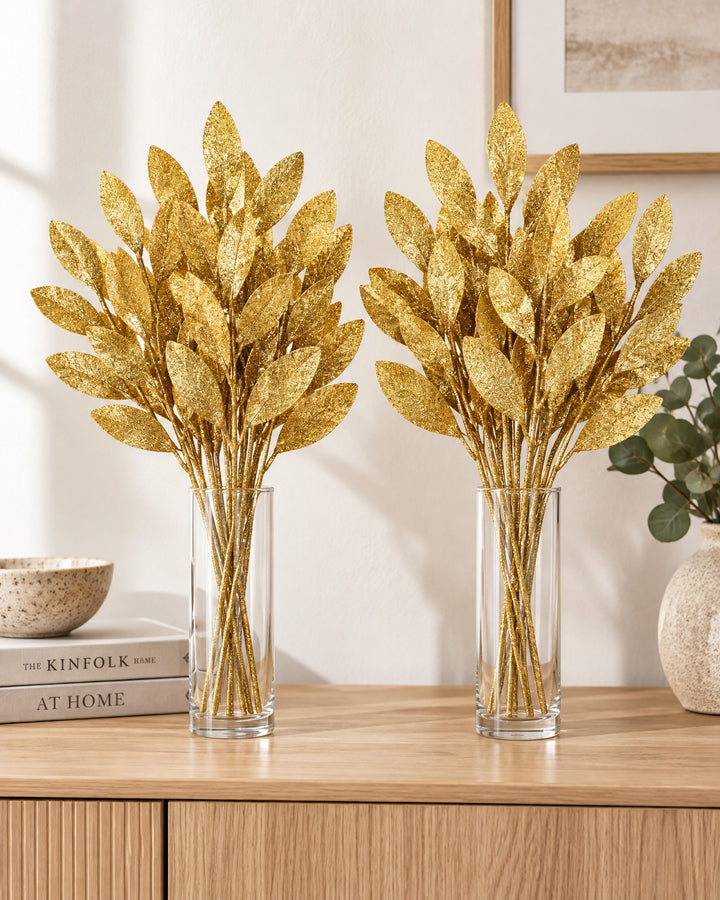 Two bunches of gold glitter leaf picks in matching glass vases on a sideboard