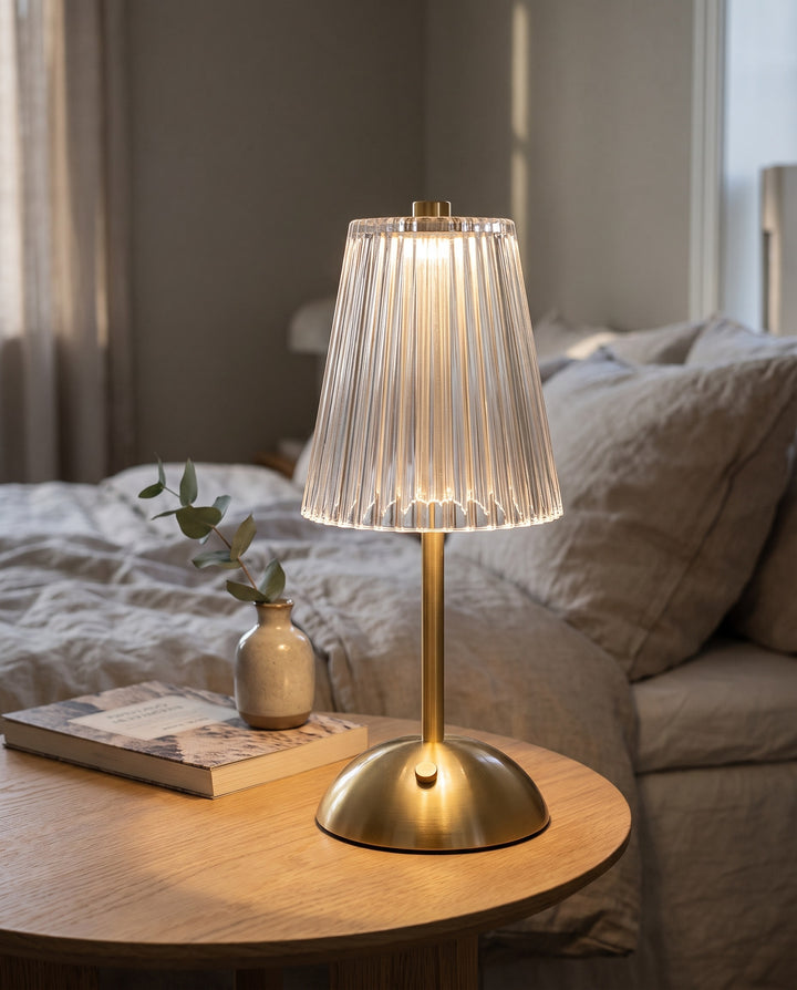 Cordless table lamp with a clear ribbed shade and gold-tone base glowing on an oak bedside table