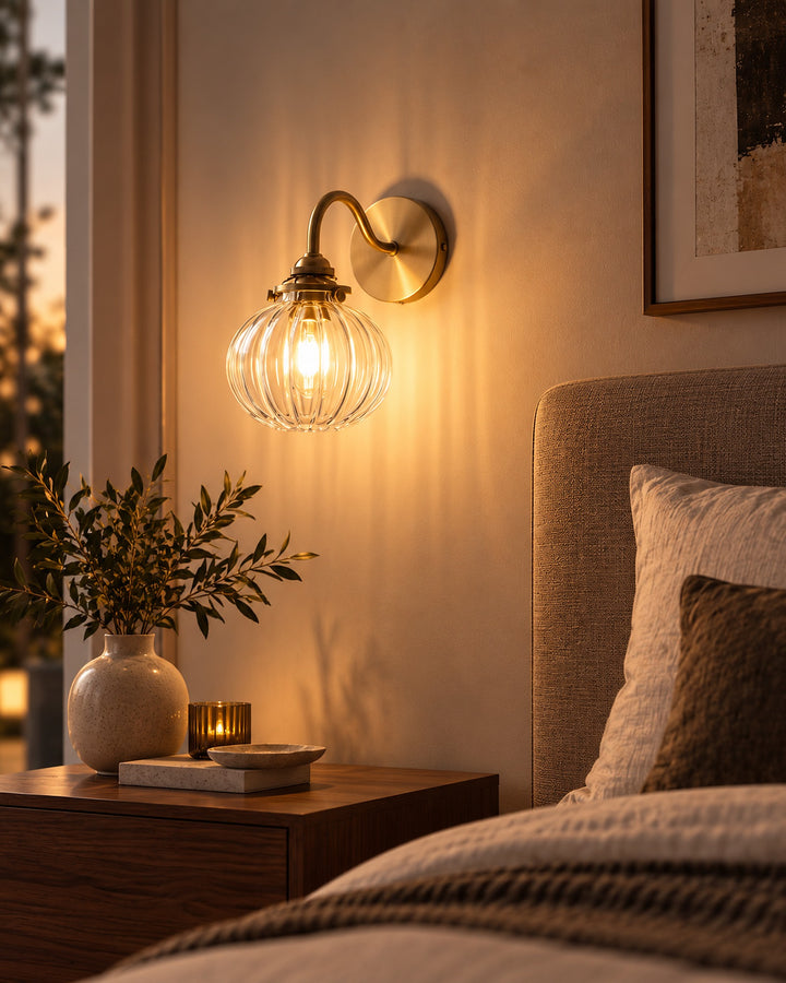 Ribbed glass globe wall sconce with a gold-tone arm, lit above a bedside table at dusk
