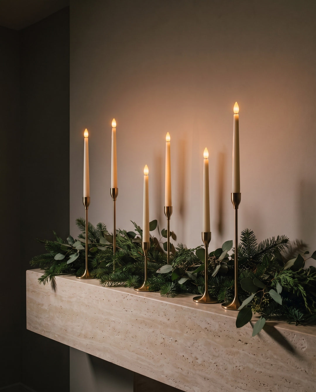 Six ivory flameless LED taper candles glowing in brass candlesticks on a travertine mantel with a fir garland